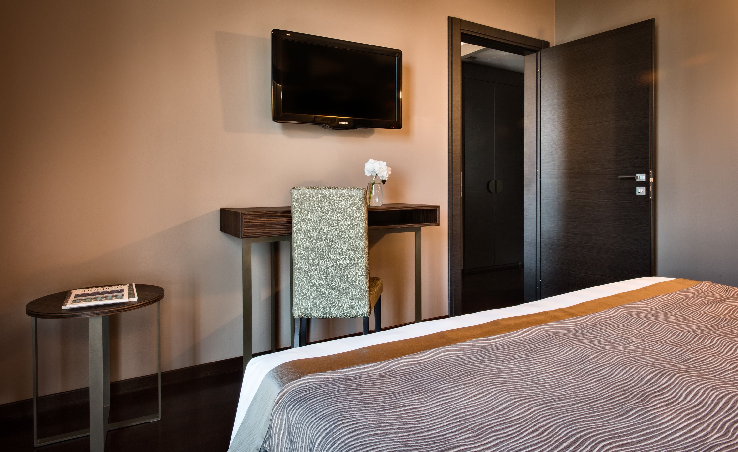 Et hotelværelse med en seng, et lille bord med en bog, et fjernsyn ophængt på væggen og en dør.