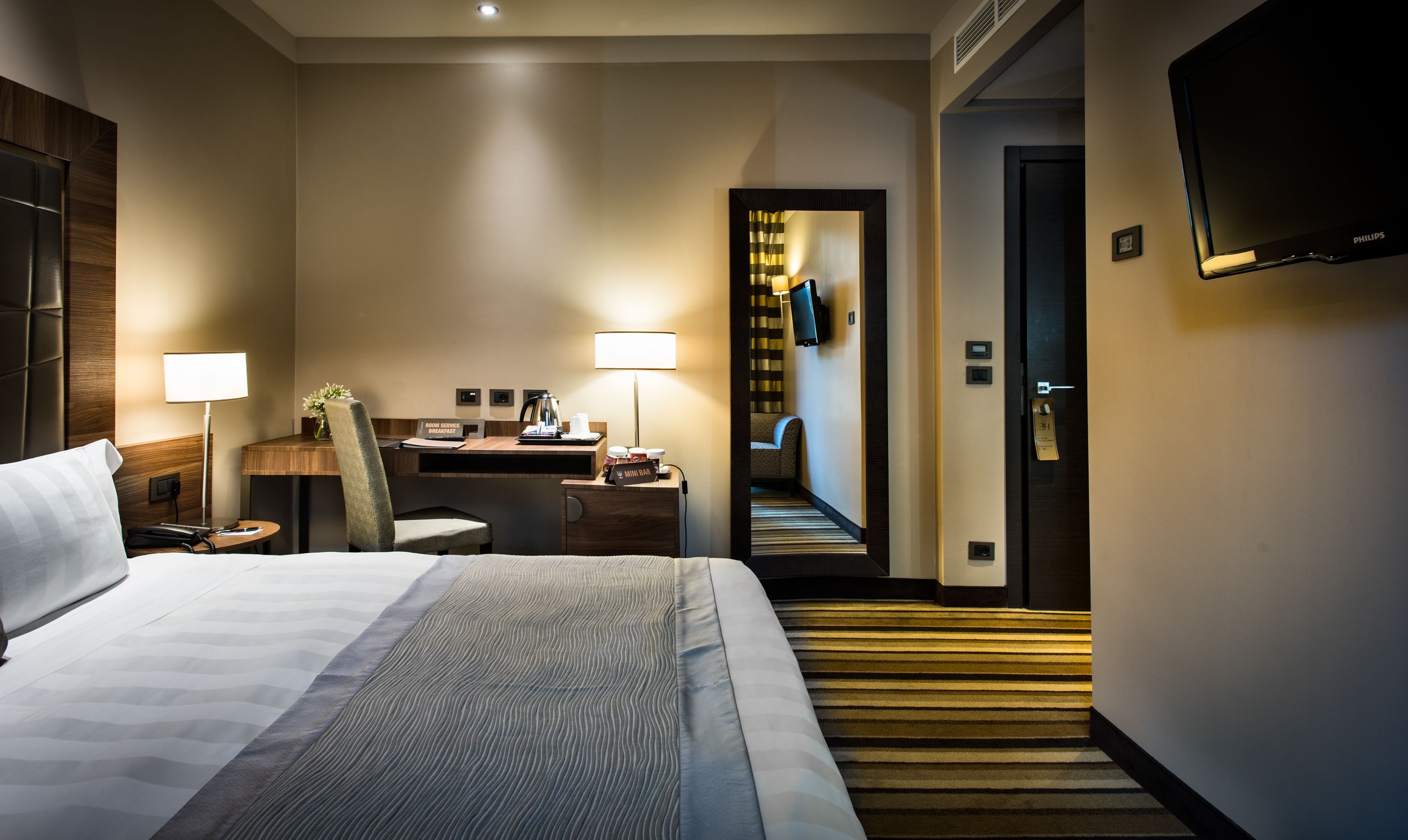 A hotel room with a bed, bedside table, lamp, and a desk with a chair. There is a mini bar and a sign indicating room service.