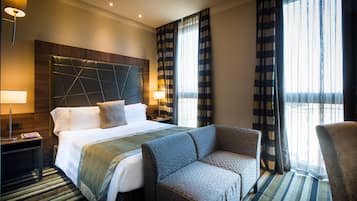 Ein Hotelzimmer mit einem großen Bett, einer Couch, einem Nachttisch, einer Lampe und Blick ins Freie durch das Fenster.