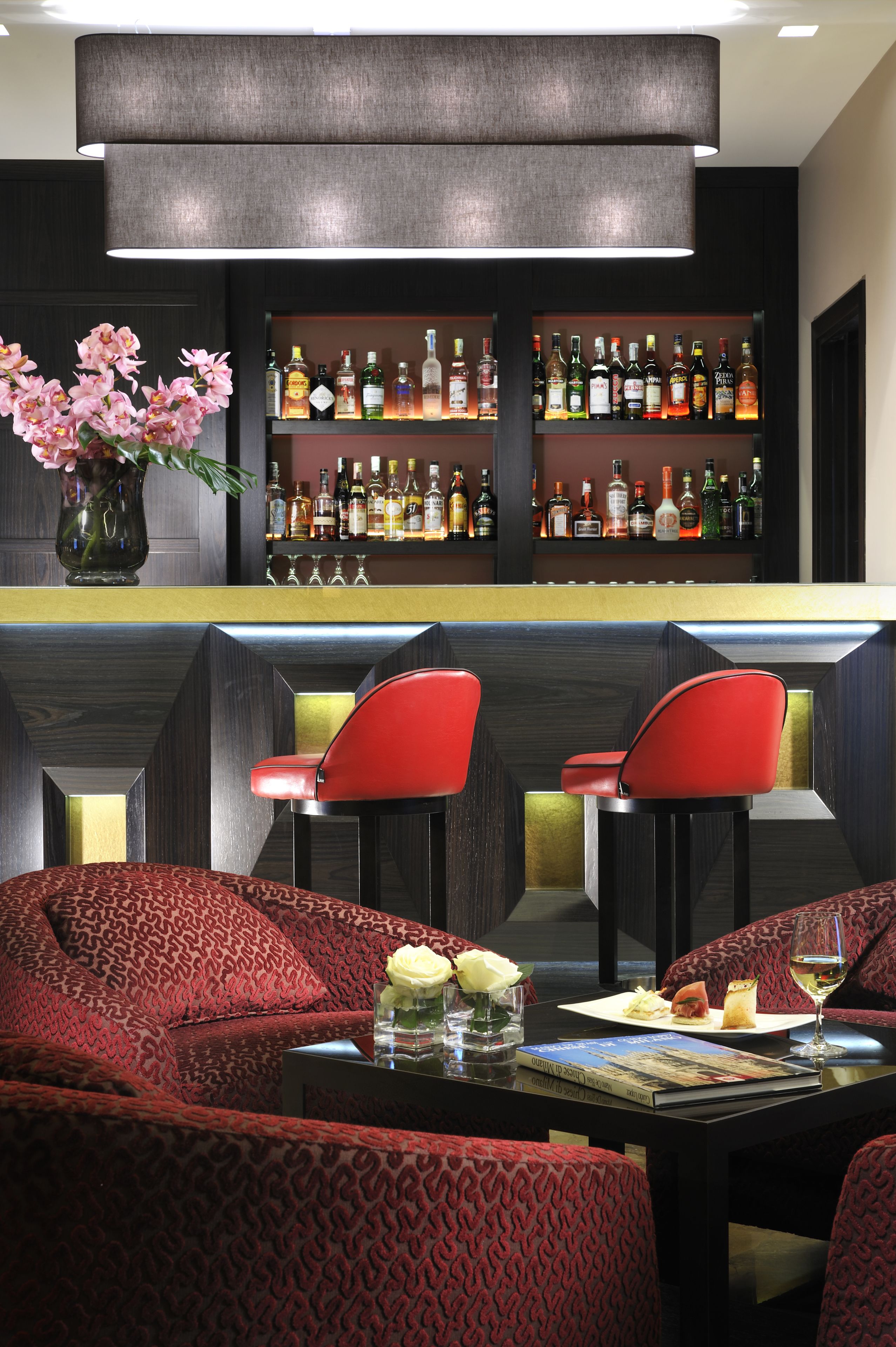 A bar area with a variety of liquor bottles, two red chairs, a table with a magazine and a glass of wine, and a vase with pink flowers.