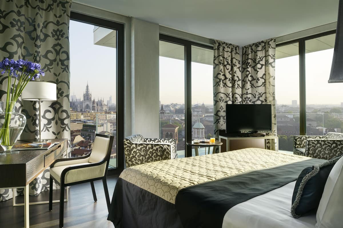 A hotel room with a large bed, a desk with a lamp, a chair, a TV, and a view of a cityscape.