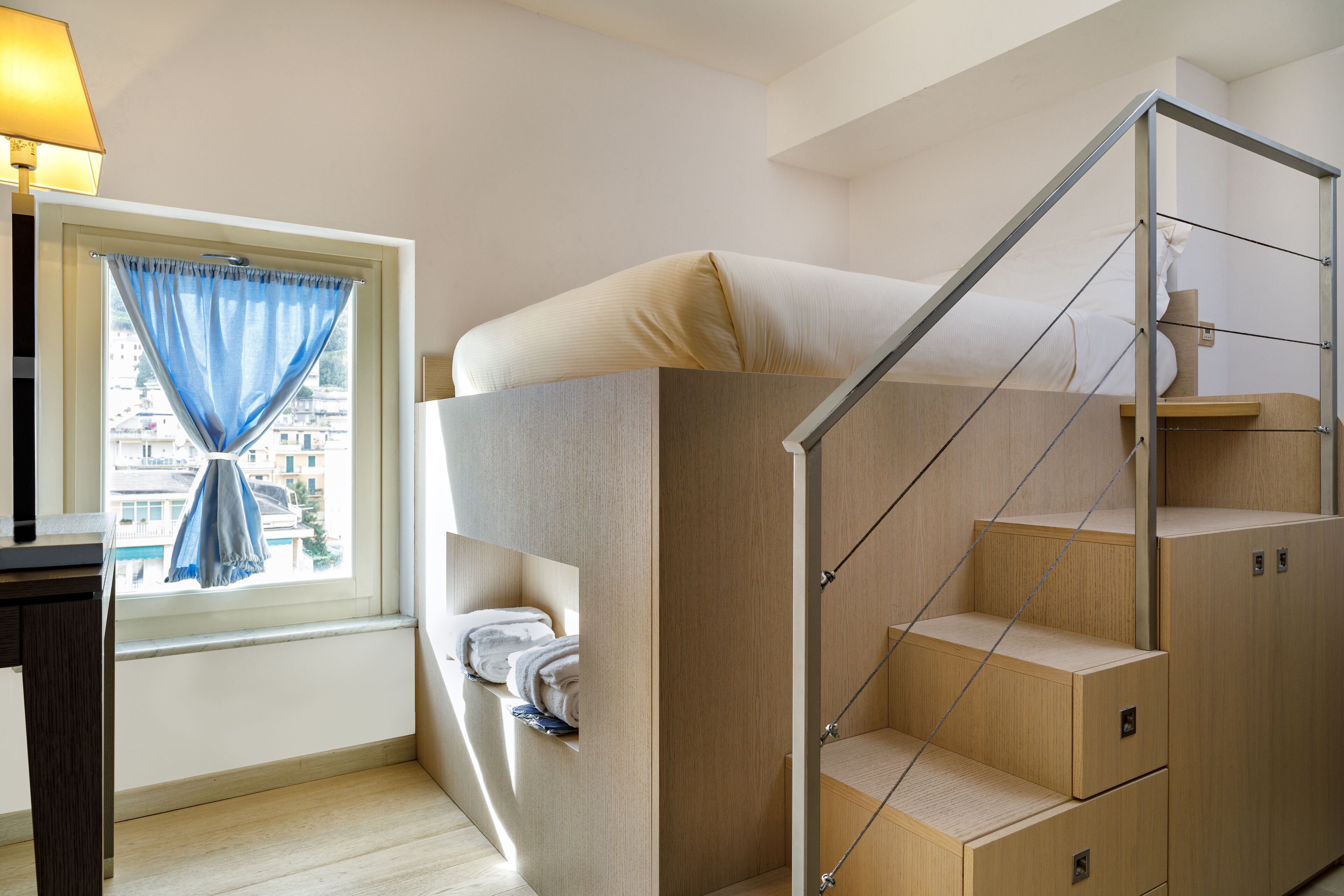 A compact room with a bunk bed, a window with curtains, a desk, and a staircase.