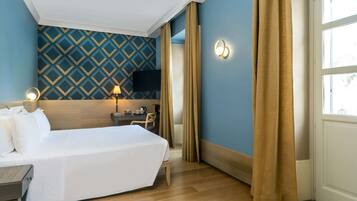 Habitación de hotel con cama, escritorio con lámpara, televisor y ventana con cortinas.
