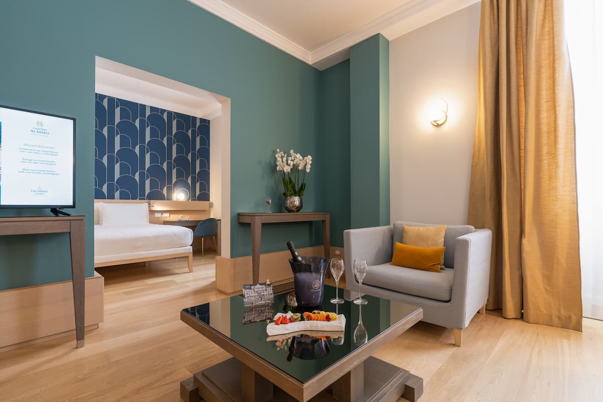 Ein modernes Hotelzimmer mit einem Bett, einem Sofa, einem Glastisch mit Speisen und Getränken, einem kleinen Tisch mit Blumenvase und einer Wandleuchte.