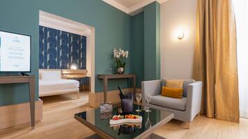Une chambre d’hôtel moderne comprenant un lit, un canapé, une table basse en verre avec de la nourriture et des boissons, une petite table avec un vase de fleurs et un luminaire fixé au mur.