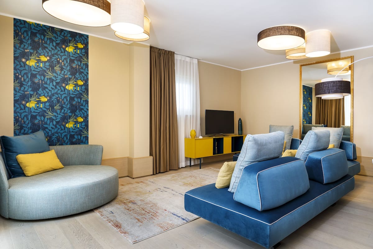 Ein modernes Wohnzimmer mit einer gebogenen grauen Couch, einer blauen Couch und einem gelben Akzentstuhl. Ein Fernseher steht auf einem gelben Schrank, es gibt einen großen Spiegel und eine Wand mit Muster.