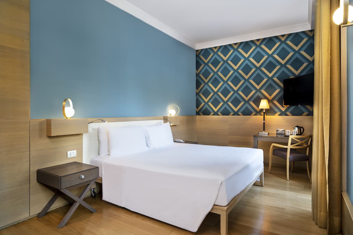 Ein Hotelzimmer mit Bett, Nachttisch, Lampe, Schreibtisch, Stuhl und Fernseher an der Wand.