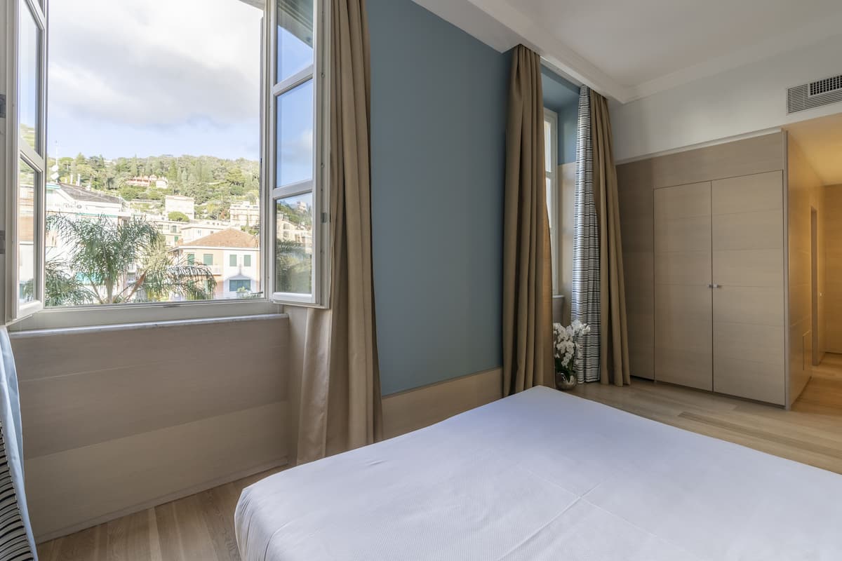 Ein Hotelzimmer mit einem großen Bett, einem Fenster mit Vorhängen, einem Kleiderschrank und Blick auf eine Hügelstadt.