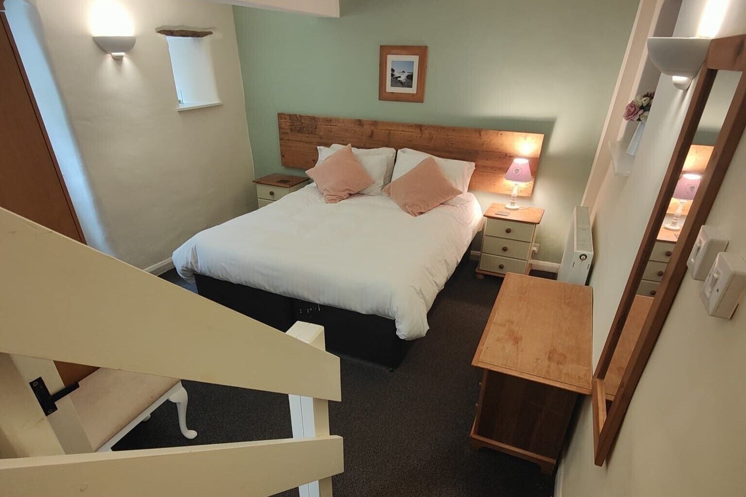 Un dormitorio con cama, mesitas de noche, cabecera de madera y una imagen enmarcada en la pared, además de una pequeña mesa con lámpara.