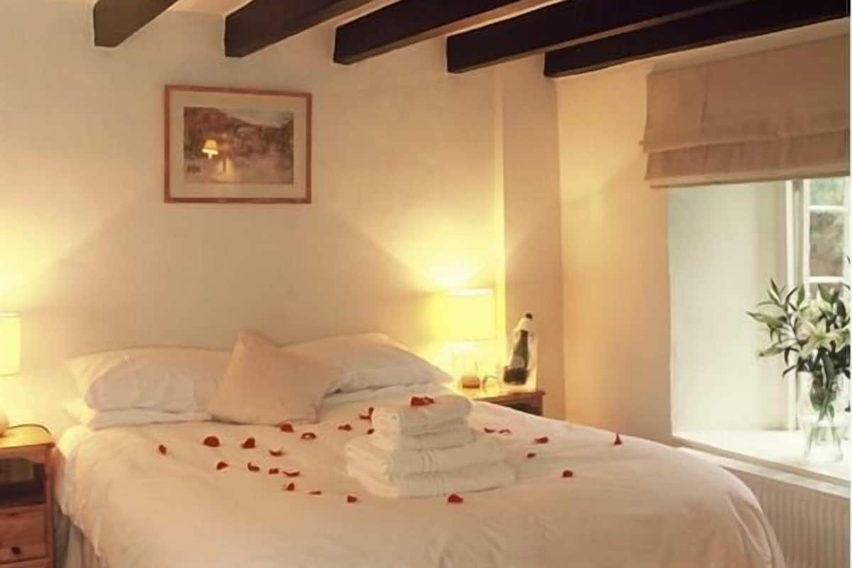 A neatly made bed with white linens and rose petals scattered on it.