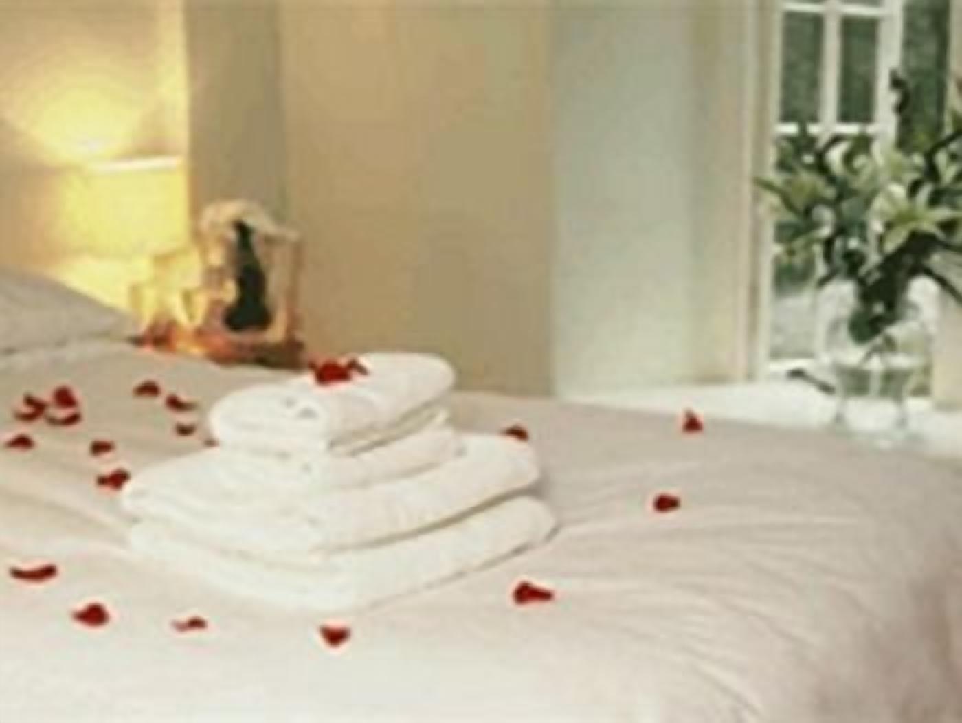 A neatly made bed with white linens and red rose petals scattered on it.