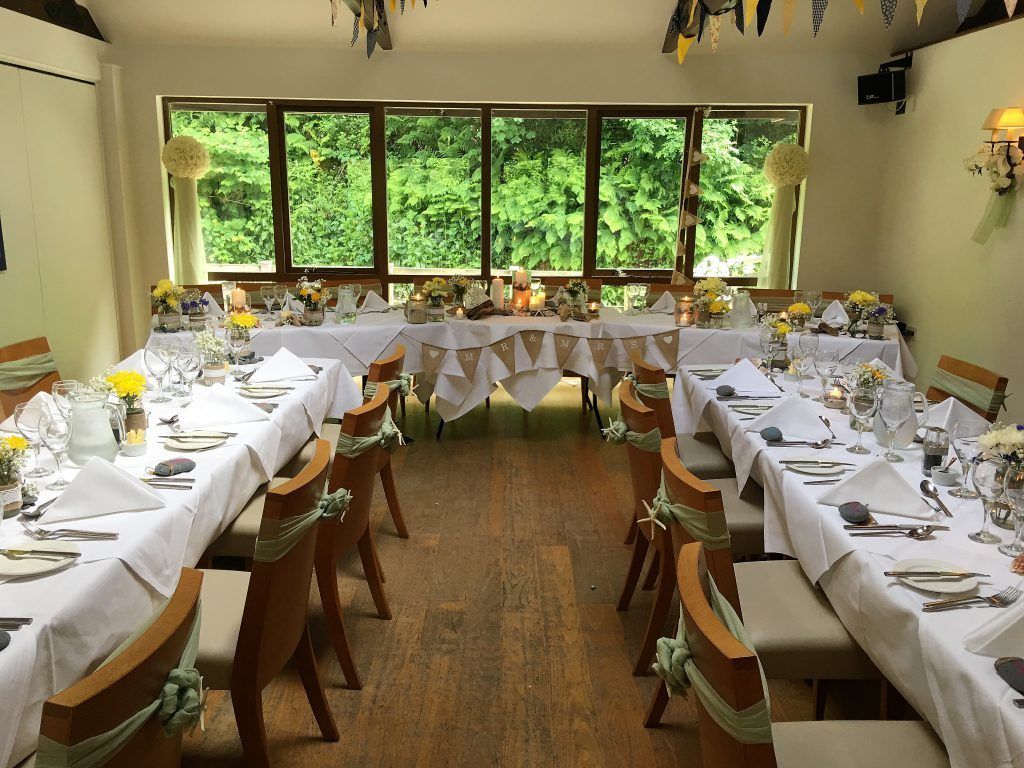 A dining area with tables set for a meal, chairs with green sashes, and large windows with a view of greenery.
