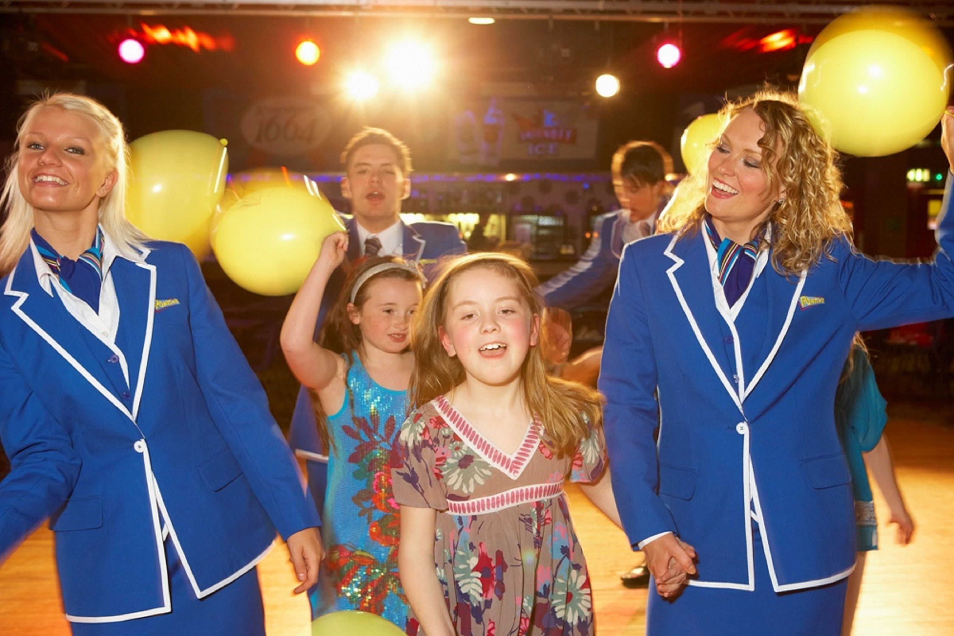 A group of people in blue uniforms holding yellow balloons in a brightly lit indoor setting.