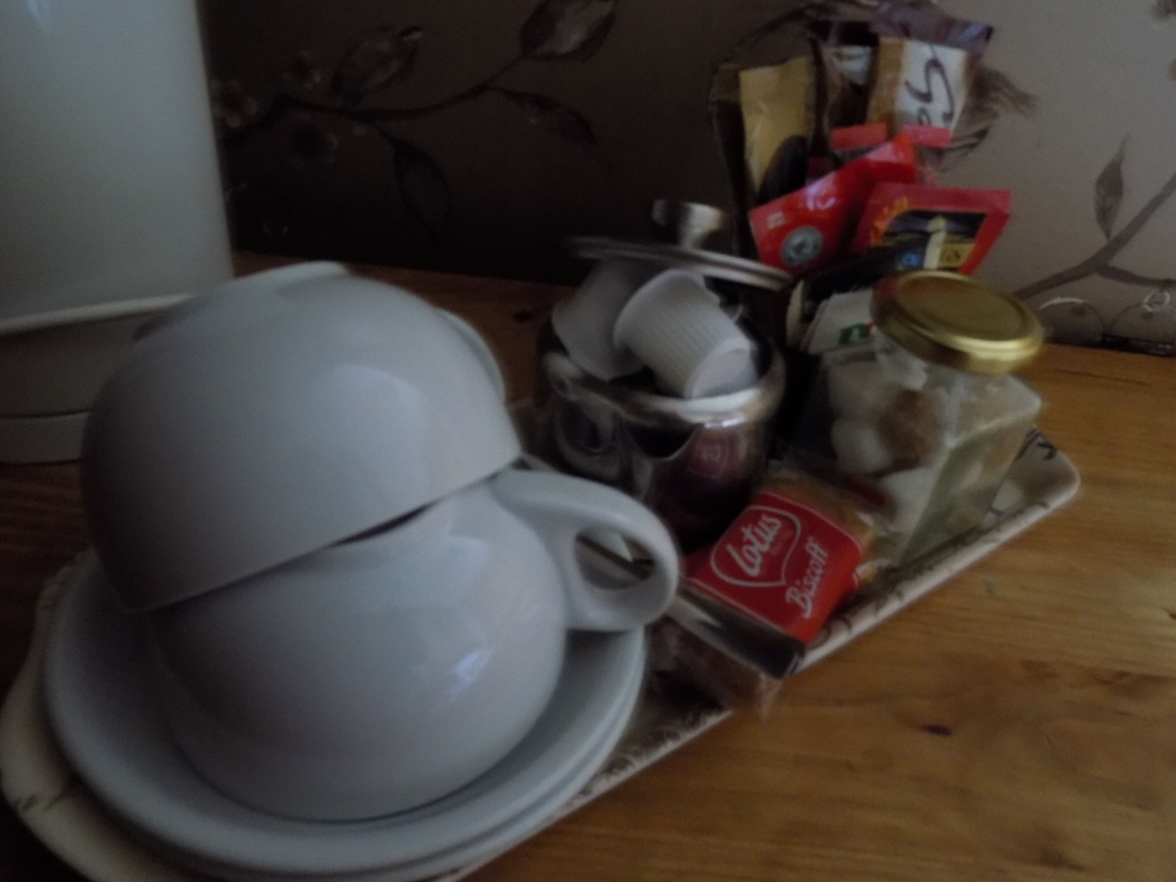 Eine weiße Keramiktasse mit Untertasse auf einem Tablett, zusammen mit einem Marmeladenglas, einer Cola-Flasche und einigen Päckchen.