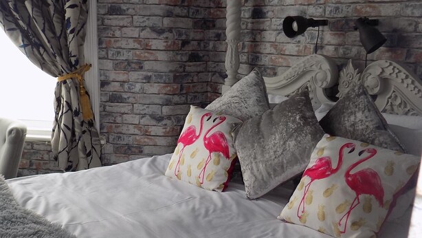 A bedroom with a brick wall, a bed with white bedding and fluffy grey throw, and decorative pillows with flamingo designs. - Driftwood Hotel (Blackpool)