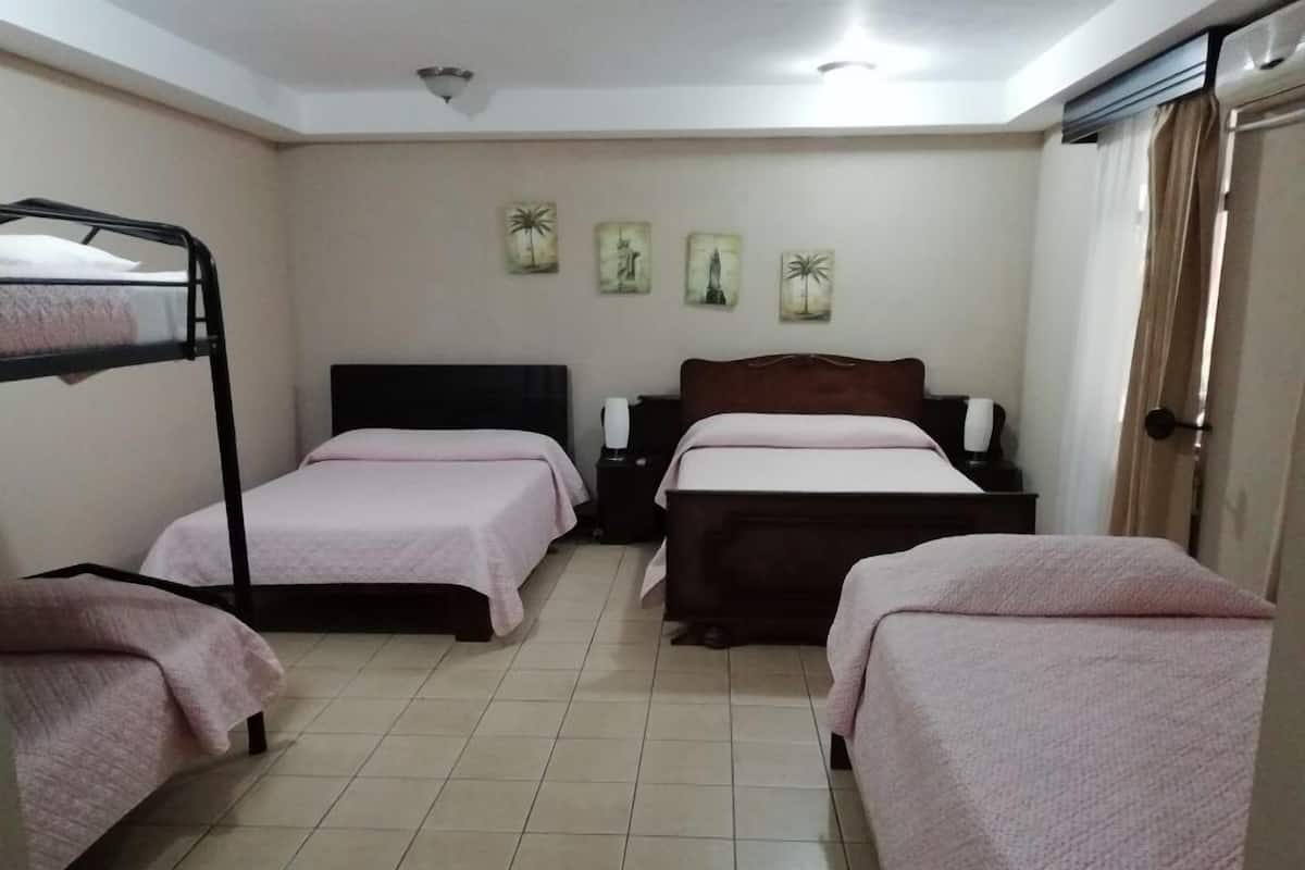 A room with two beds, one with a black frame and one with a wooden frame, both with pink bedding. There are three wall-mounted paintings and a ceiling light.