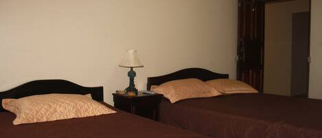 A bedroom with two beds, a nightstand with a lamp, and a door leading to another room.