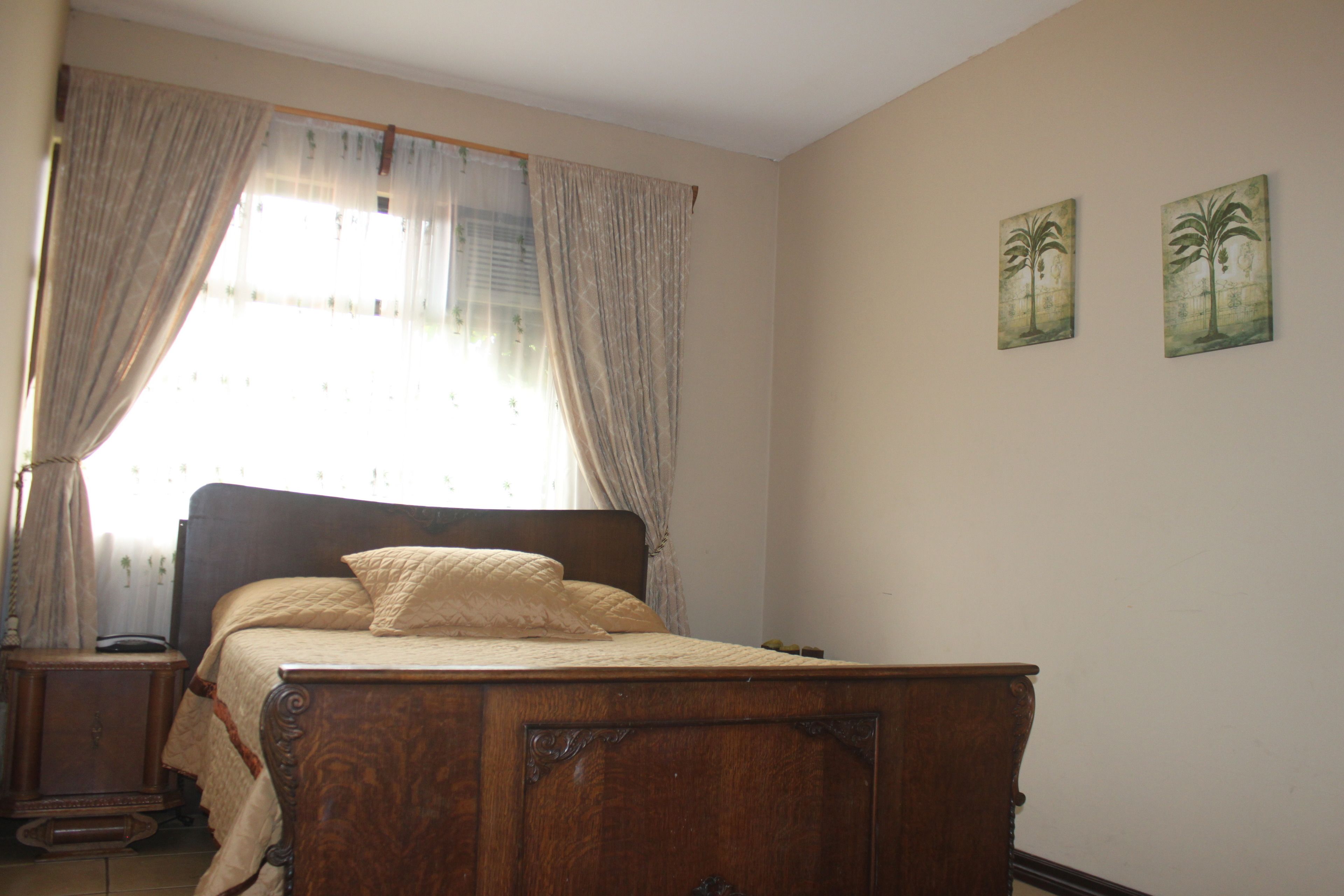 A bedroom with a wooden bed, a nightstand, a window with sheer curtains, and two wall-mounted paintings.
