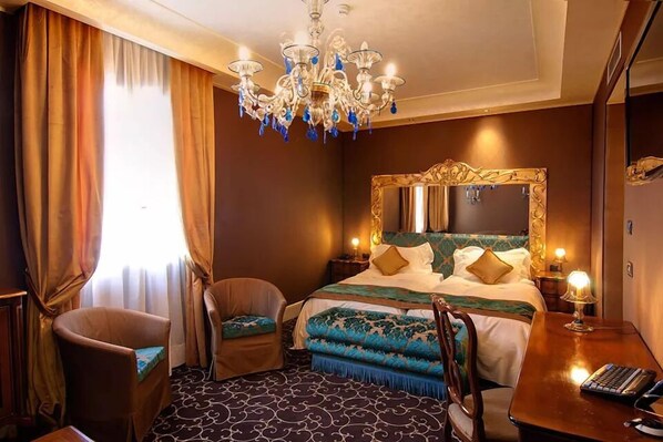 A hotel room with a large bed, a chandelier, a desk with a lamp, and a chair. - Pesaro Palace (Venice)