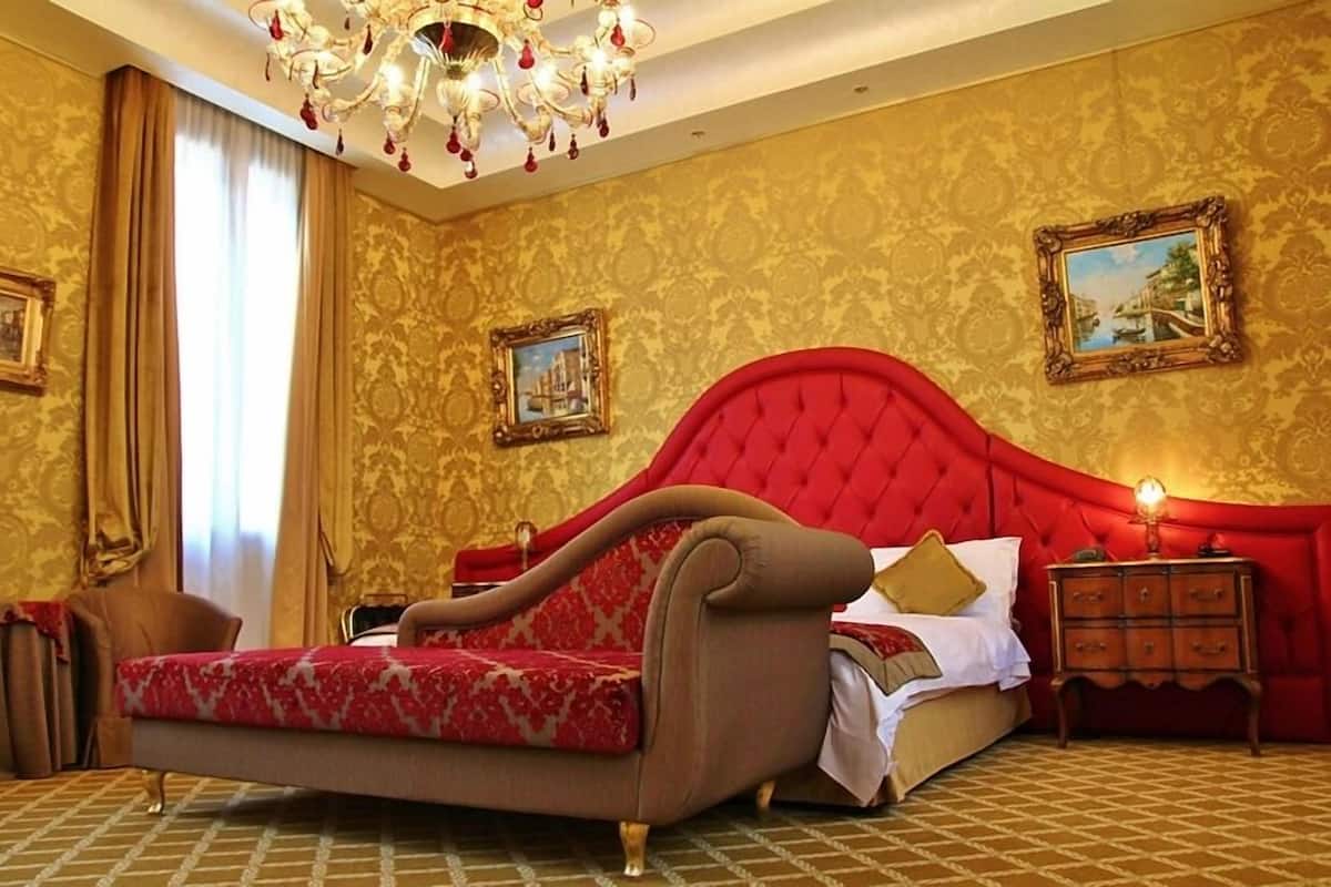 A luxurious bedroom with a red upholstered bed, a chandelier, and ornate wallpaper.