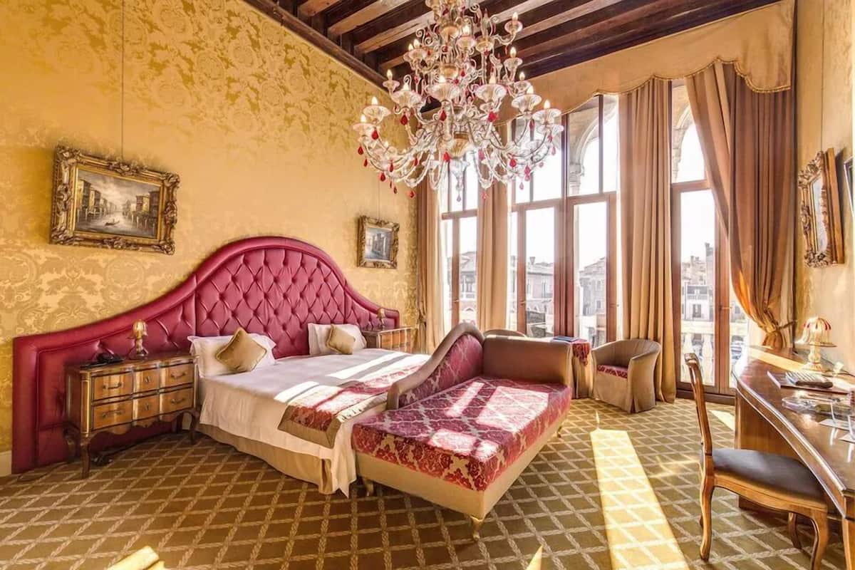 A room with a large bed, a desk, and a chair. There are two windows with curtains, a chandelier, and a patterned floor.