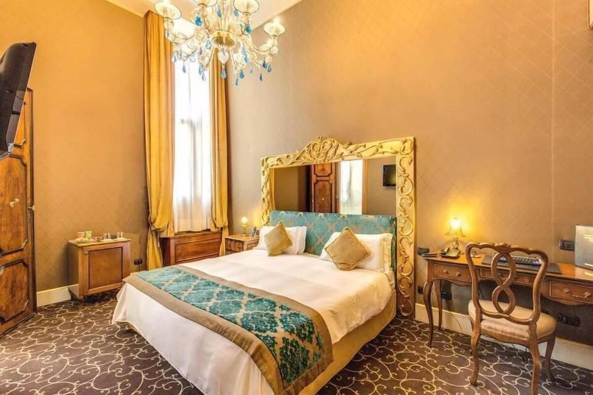 A hotel room with a large bed, a desk, a chair, a wardrobe, and a chandelier.