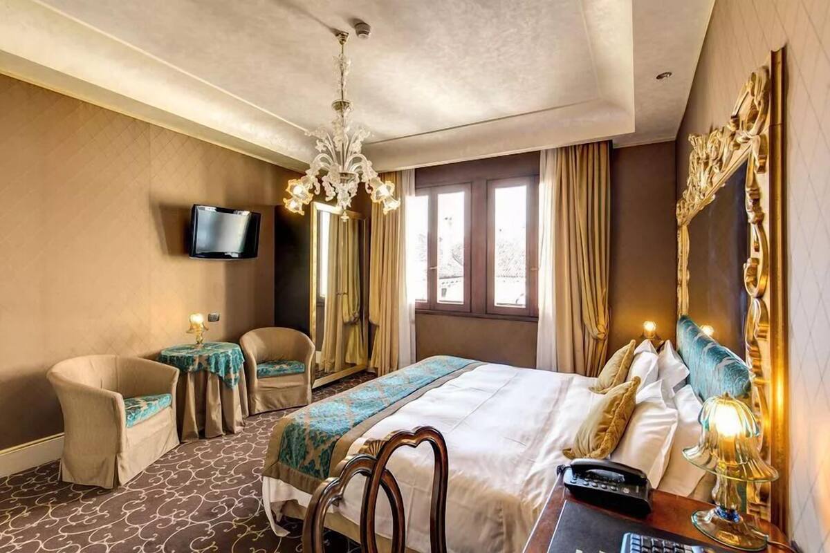 A hotel room with a large bed, a desk with a telephone, a chair, a television mounted on the wall, and a chandelier.