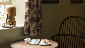 Ein runder Holztisch mit einer Kaffeetasse, einer Schüssel Sahne und einer Speisekarte. Ein karierter Stuhl steht vor dem Tisch.