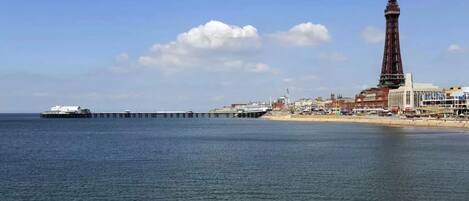 Una escena costera con un muelle que se adentra en el mar, una torre destacada y una playa con edificios al fondo.
