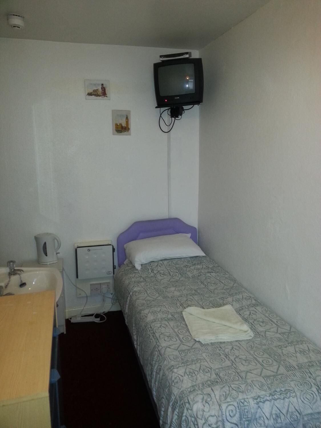 A small, simple room with a single bed, a bedside table, a television mounted on the wall, and a small cabinet.