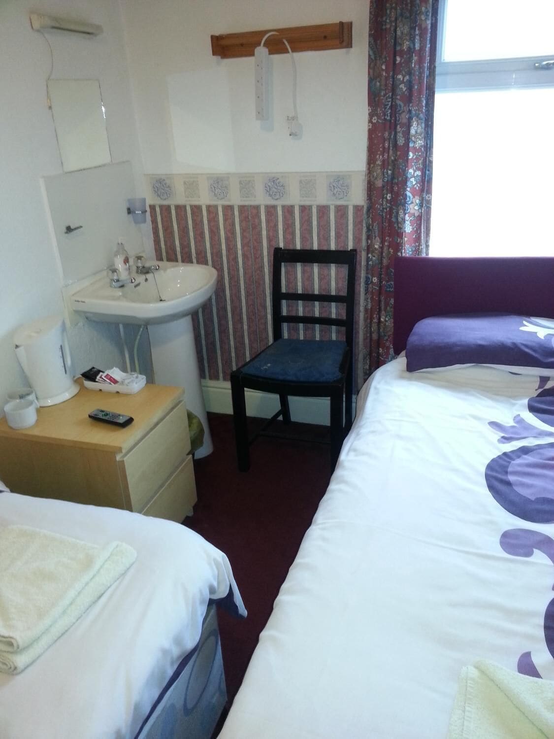 Habitación de hotel con dos camas, un lavamanos pequeño, una silla y una ventana con cortinas.