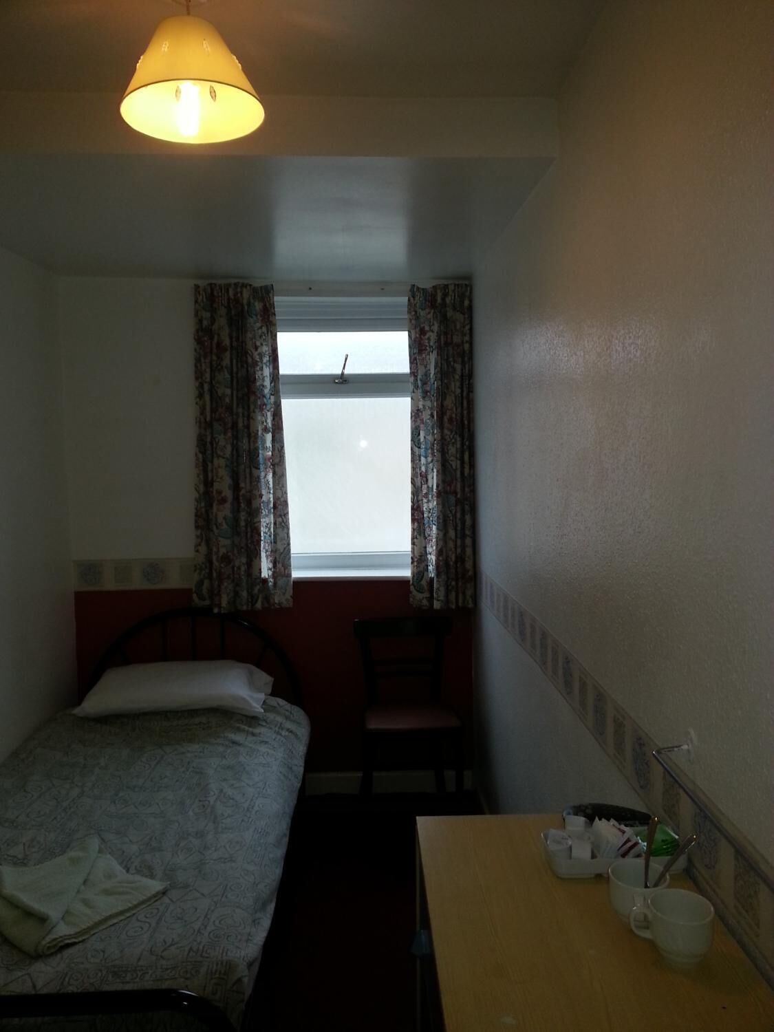 A small, dimly lit room with a single bed, a chair, a desk with a cup and a book, and a window with curtains.