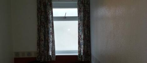 A small, dimly lit room with a single bed, a chair, a desk with a cup and a book, and a window with curtains.