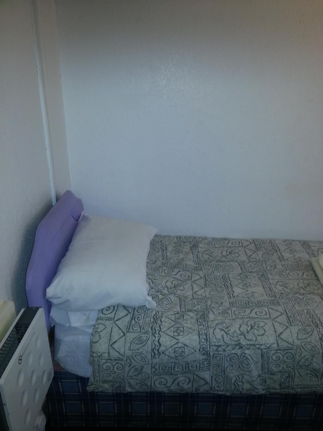A small, single-bed room with a patterned bedspread, a purple pillow, and a white pillow.