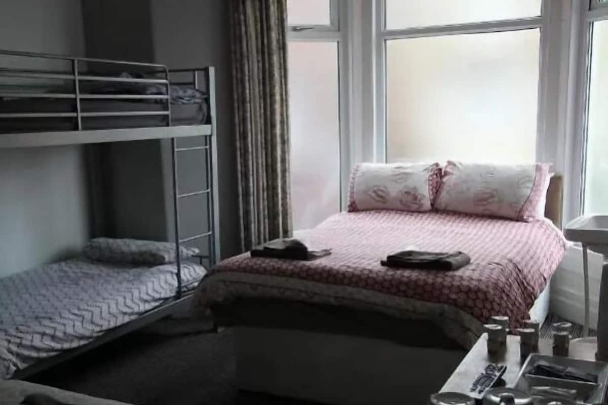 A bedroom with a bunk bed, a single bed, a desk with a chair, and a window with curtains.