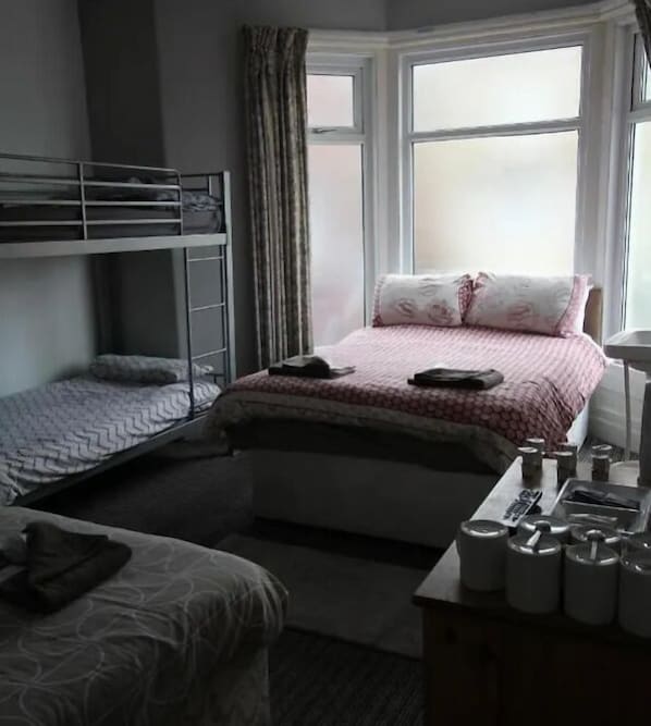 A bedroom with a bunk bed, a single bed, a desk with a chair, and a window with curtains. - Jesmond International Hotel (Blackpool)