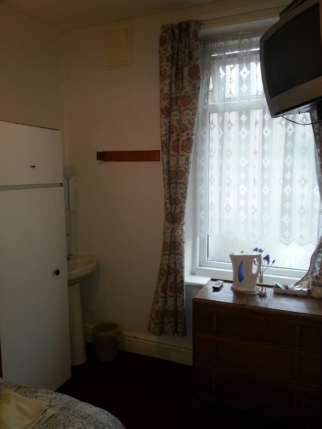A small, dimly lit room with a toilet, a bed, a television, and a window with patterned curtains.