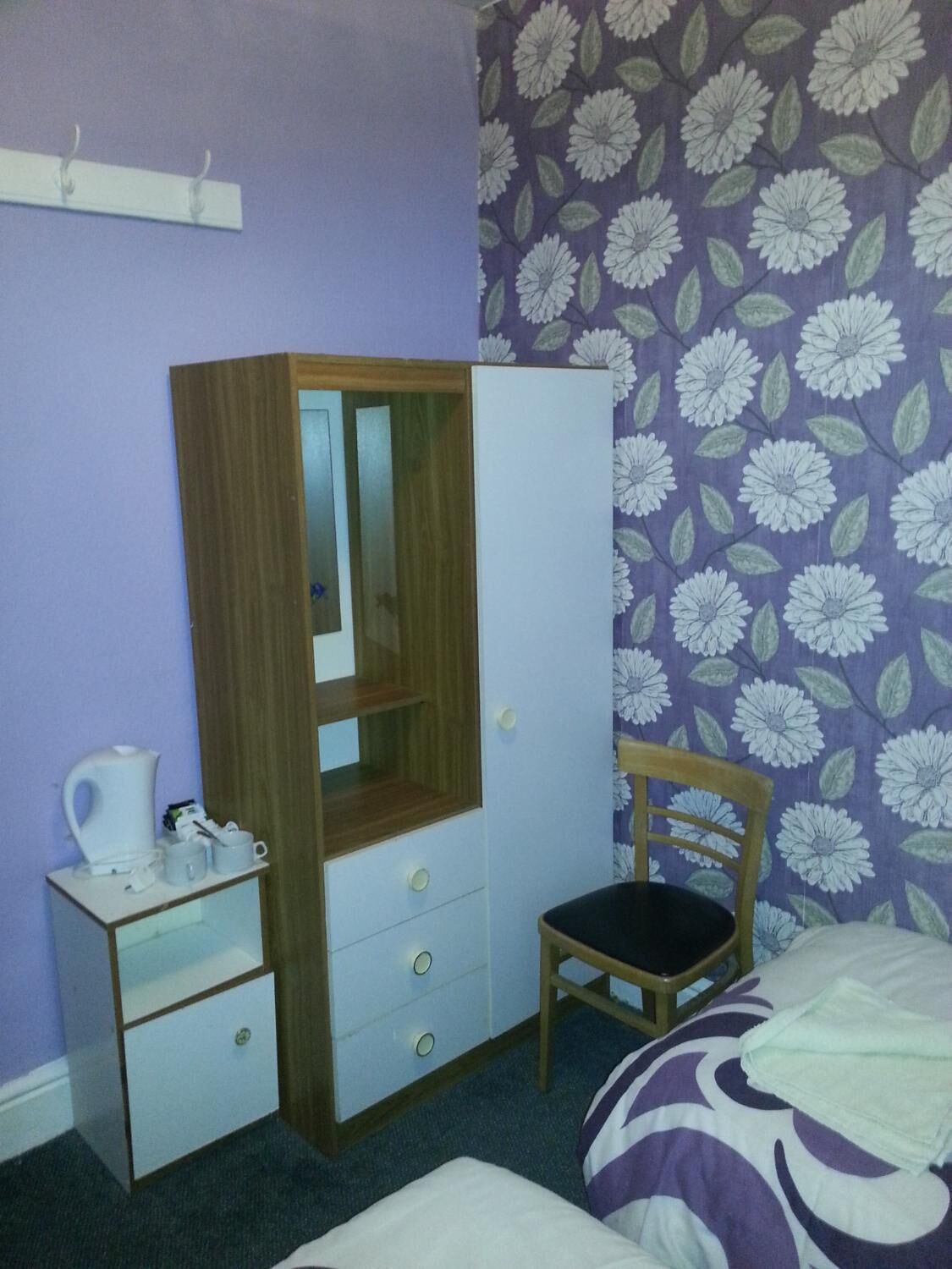A bedroom with a wooden wardrobe, a chair, a bed with a purple and white patterned bedspread, and a floral wallpaper.
