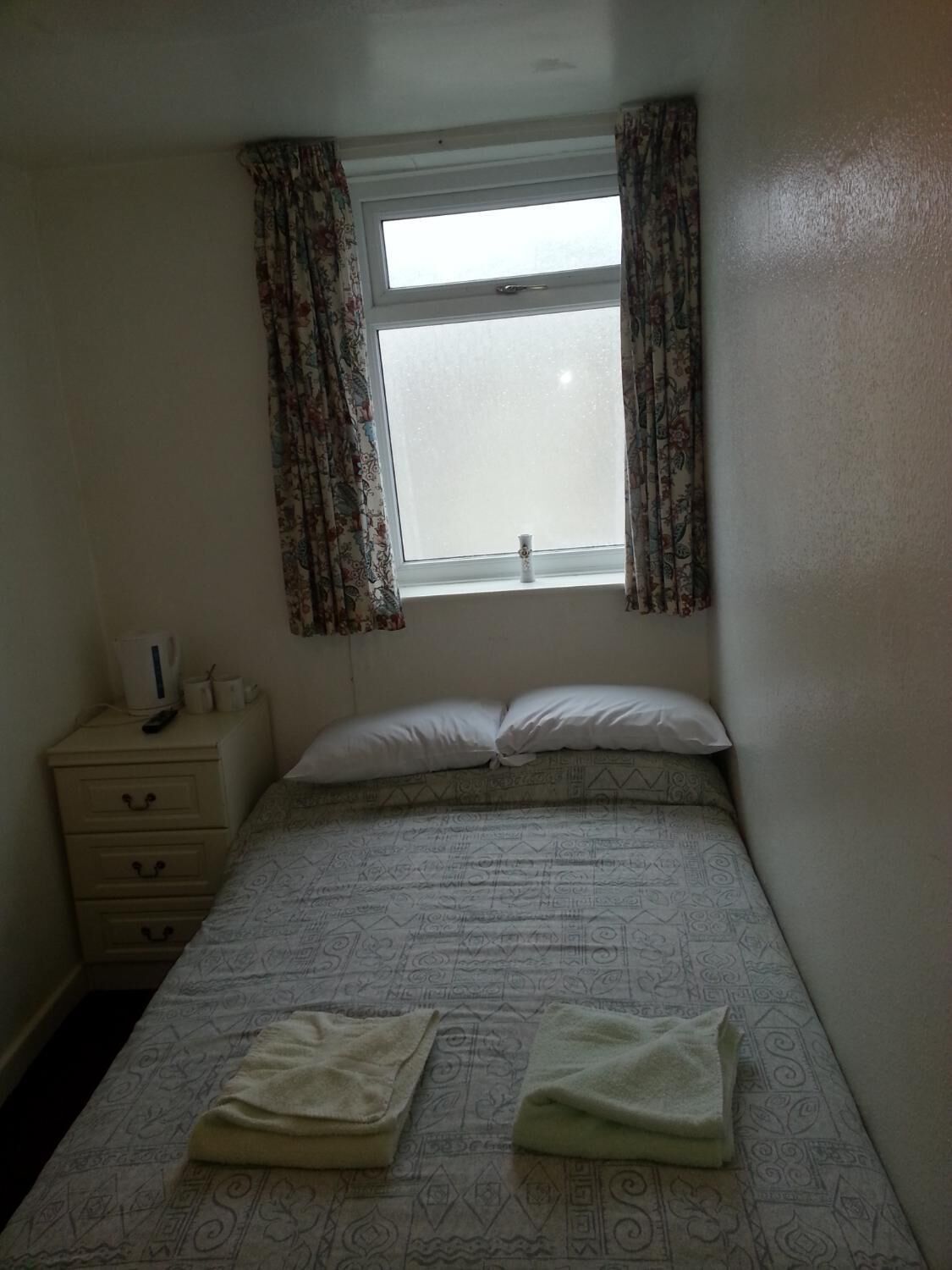 A small, single-bed room with a window, a bedside table, and a nightstand.