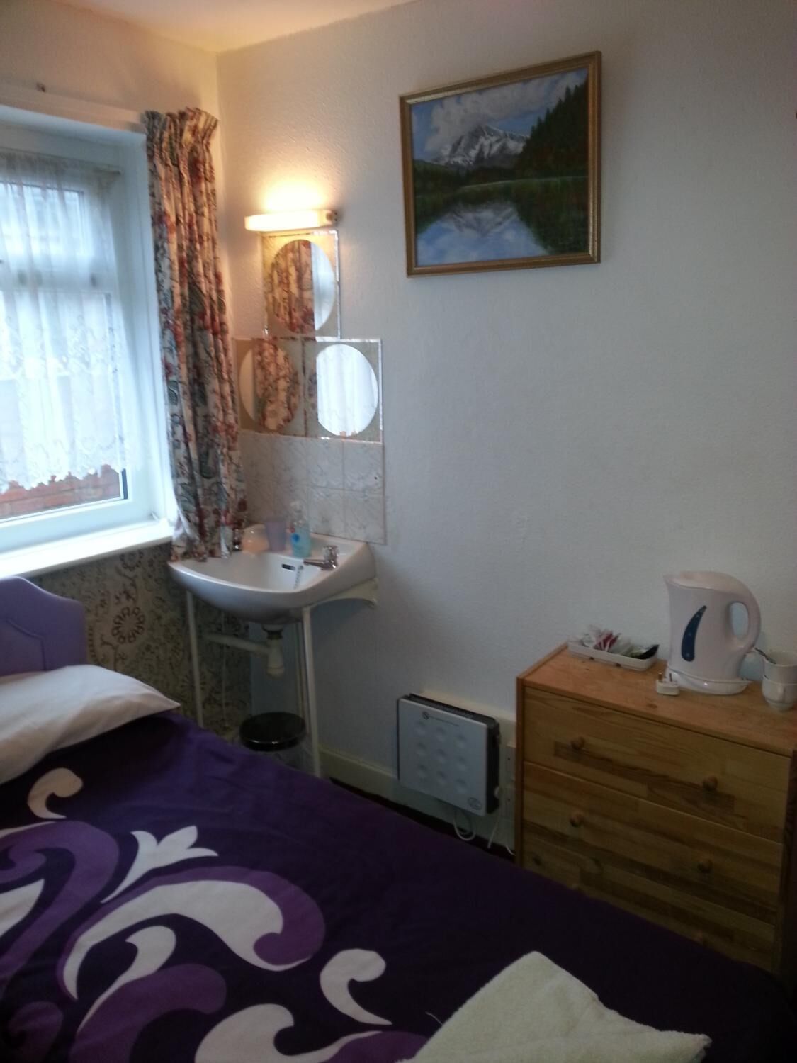 Une chambre à coucher comprenant un lit, une table de chevet avec une bouilloire, une commode, une fenêtre avec des rideaux, un lavabo et un tableau accroché au mur.