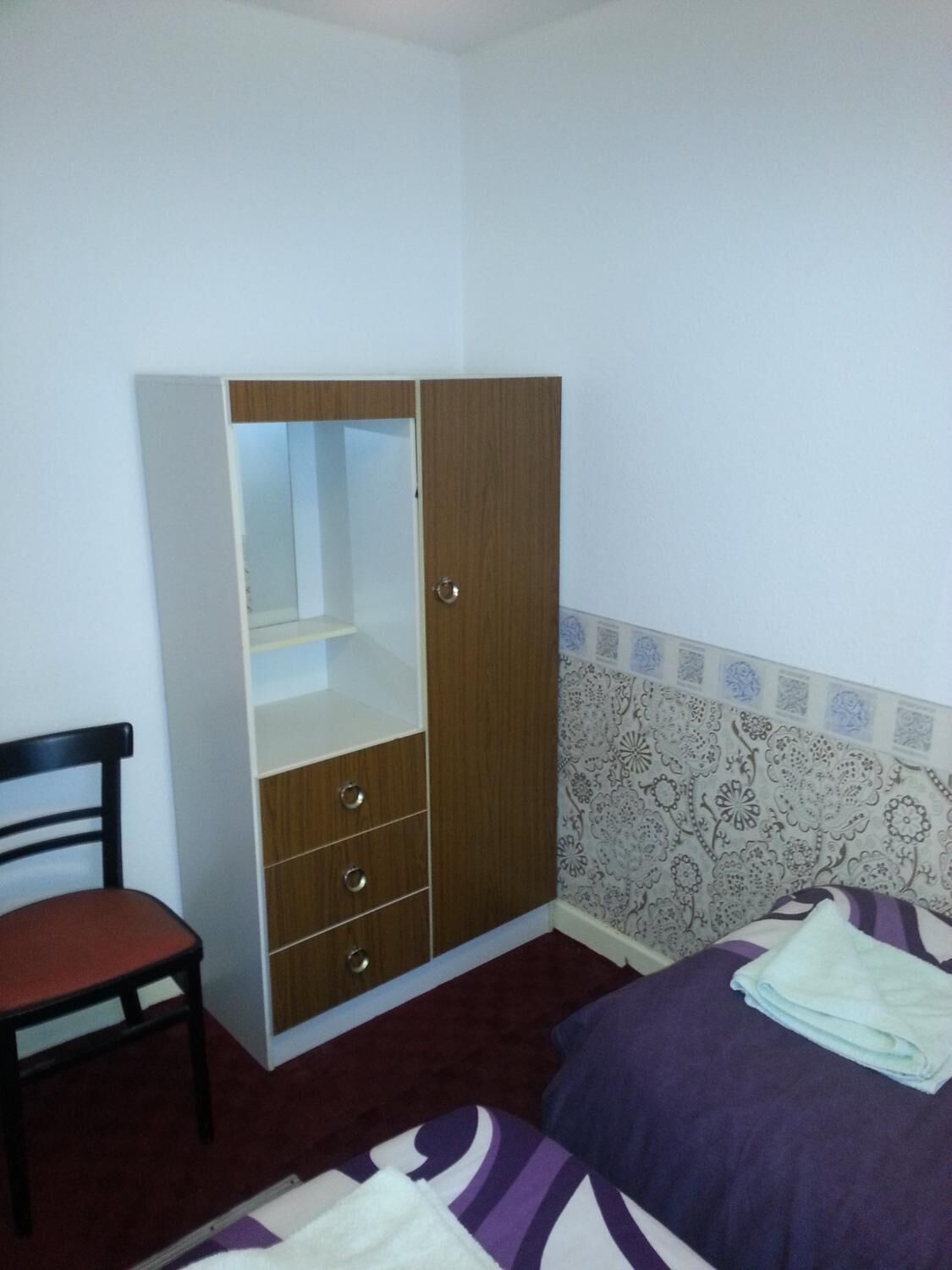 Een slaapkamer met een bed, een houten garderobe met spiegel, een stoel en behang met een patroon.
