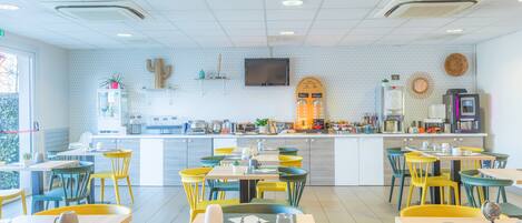 Una cafetería moderna con un mostrador central, varias zonas de estar con sillas coloridas y una cocina visible al fondo.