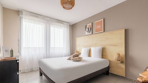A hotel room with a bed, a desk, a chair, a window with curtains, and a wooden headboard. - Appart'City Confort Toulouse Aéroport Cornebarrieu (Cornebarrieu)