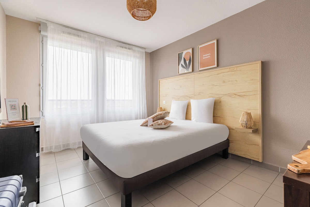 A hotel room with a bed, a desk, a chair, a window with curtains, and a wooden headboard.