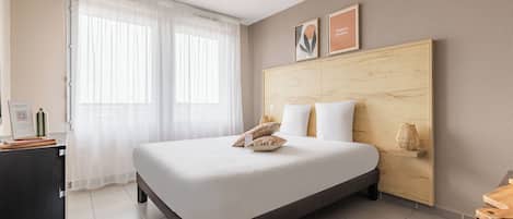 A hotel room with a bed, a desk, a chair, a window with curtains, and a wooden headboard.