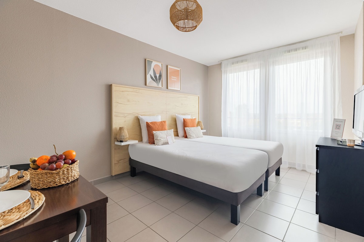 A hotel room with a bed, a desk, a basket of fruit, and a window with curtains.