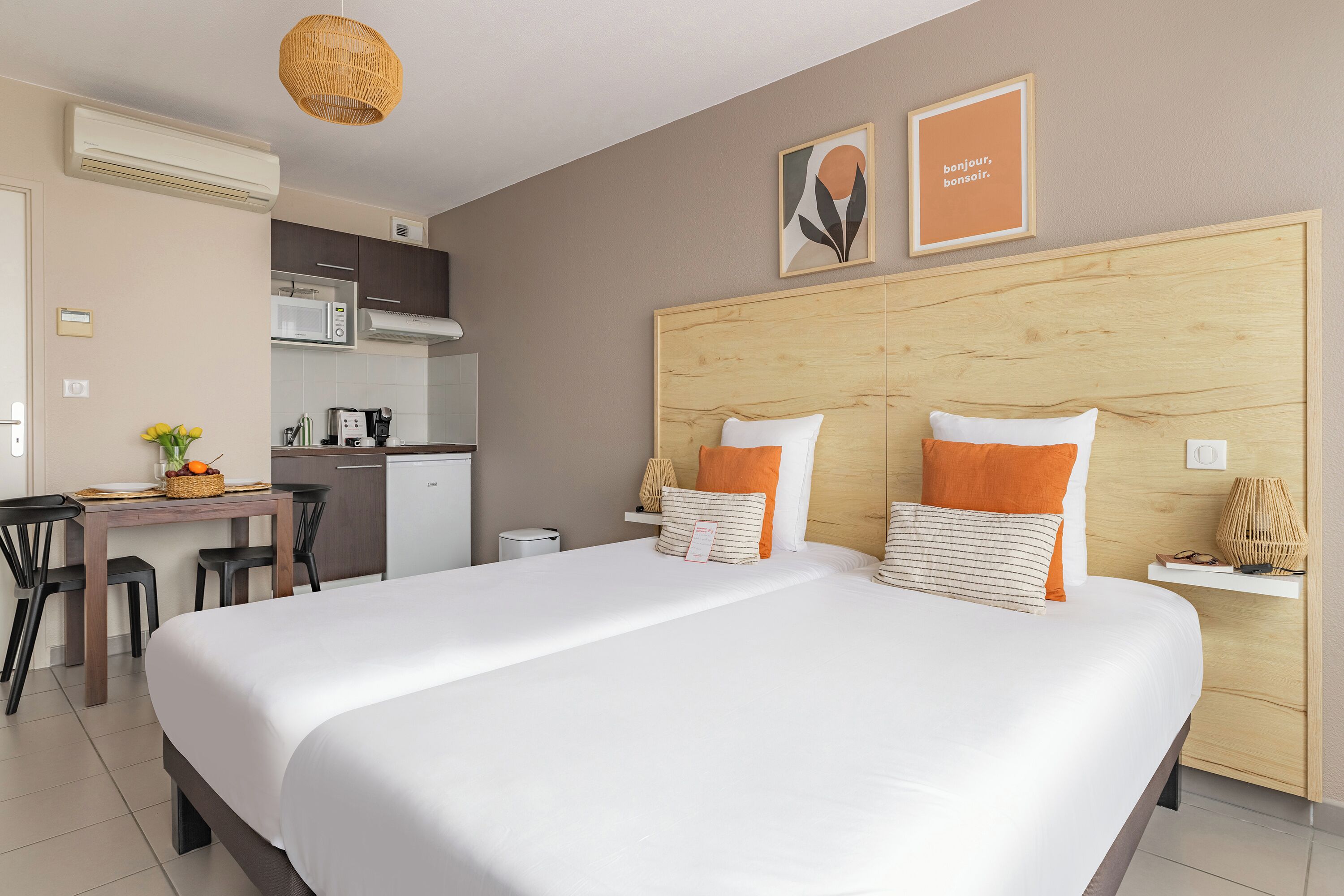 Quarto de hotel com uma cama grande, cabeceira de madeira, escrivaninha com cadeira, cozinha compacta e ar-condicionado fixado na parede.