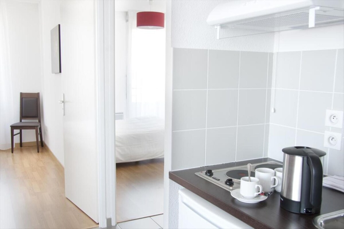 A hotel room with a bed, a chair, a dining area with a kettle, cups, and a stove.