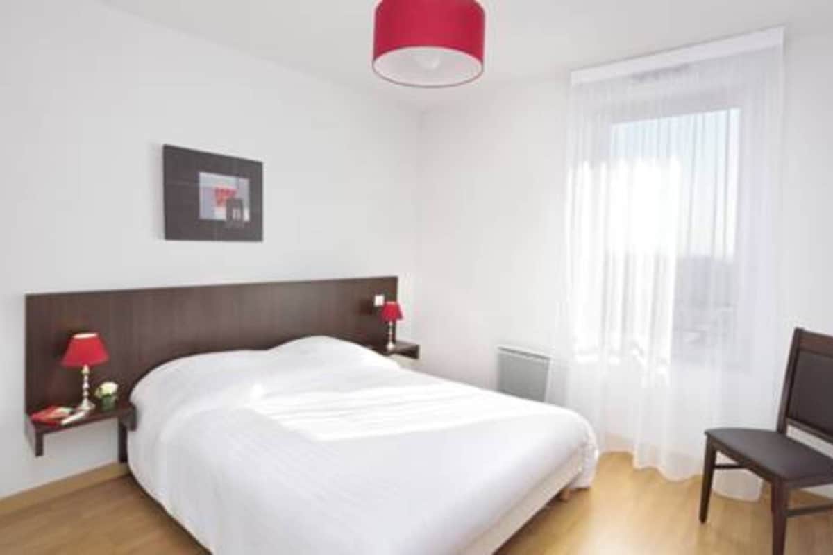 Una habitación de hotel con una cama, una mesita de noche con lámpara, una silla, una ventana con cortinas translúcidas y un cuadro en la pared.