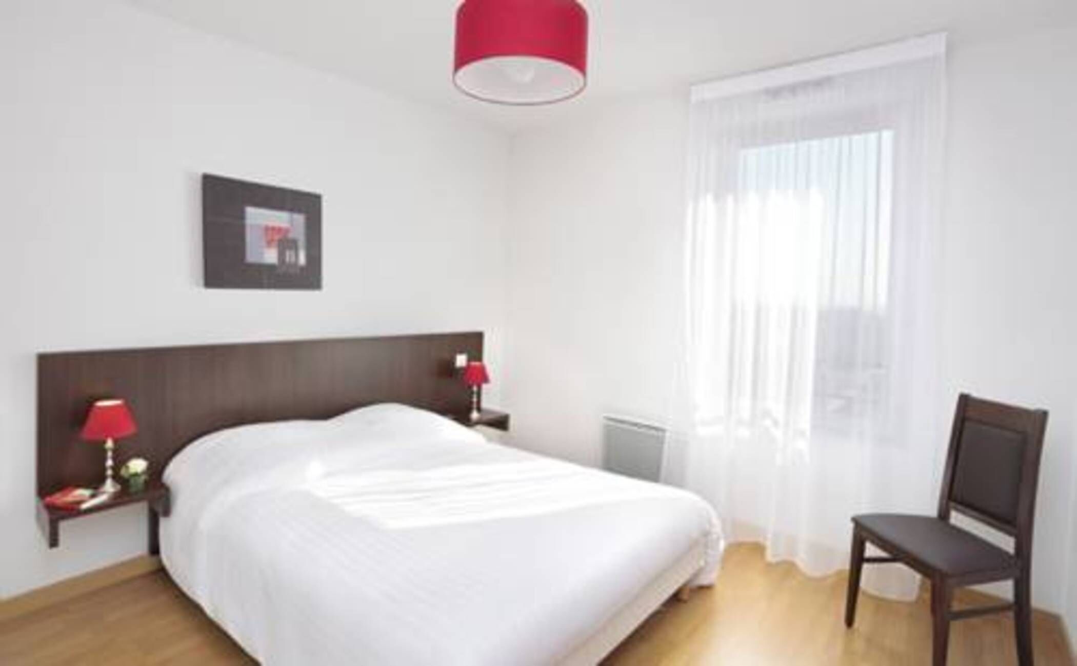 Een hotelkamer met een bed, een nachtkastje met een lamp, een stoel, een raam met dunne gordijnen en een foto aan de muur.